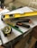 A collection of tools including a yellow level, a measuring tape, a utility knife, a green pencil, and a black marker are placed on top of some stacked papers and a plastic box. There is additional construction equipment in the background.
