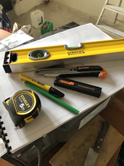 A collection of tools including a yellow level, a measuring tape, a utility knife, a green pencil, and a black marker are placed on top of some stacked papers and a plastic box. There is additional construction equipment in the background.
