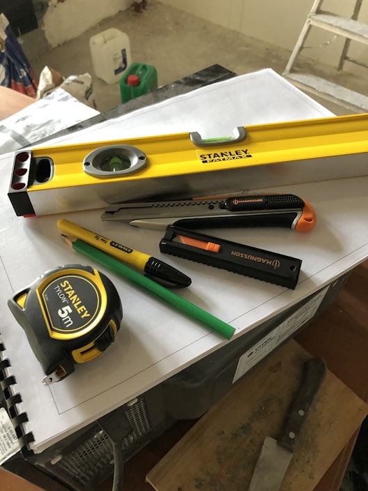 A collection of tools including a yellow level, a measuring tape, a utility knife, a green pencil, and a black marker are placed on top of some stacked papers and a plastic box. There is additional construction equipment in the background.