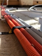 Several corrugated cables in orange and gray are neatly bound together with a black strap, lying on a wooden or concrete surface with sunlight casting shadows across them.