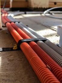 Several corrugated cables in orange and gray are neatly bound together with a black strap, lying on a wooden or concrete surface with sunlight casting shadows across them.