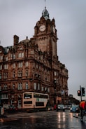 A large, ornate stone building with a clock tower. The architecture features intricate detailing and arched windows. The foreground shows a busy street with a double-decker bus, cars, and a person in a red jacket. Flags, including a Scottish flag, are hanging from the building.