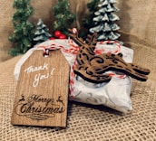 A rustic wooden tag inscribed with 'Thank You!' and 'Merry Christmas' is attached to a wrapped gift with decorative twine. A wooden reindeer ornament accompanies the tag, and the background features small, snowy pine trees on burlap fabric.