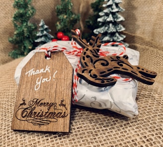A rustic wooden tag inscribed with 'Thank You!' and 'Merry Christmas' is attached to a wrapped gift with decorative twine. A wooden reindeer ornament accompanies the tag, and the background features small, snowy pine trees on burlap fabric.