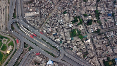 An aerial view of a densely populated urban area featuring a complex network of roads and highways. The image includes multiple lanes, overpasses, and a roundabout. Buildings are tightly packed, and patches of greenery can be seen scattered throughout.
