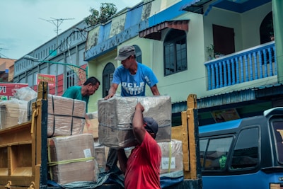 Three individuals are unloading large cardboard boxes from a truck onto the street in front of a row of colorful buildings. They appear to be working together to carefully handle the packages, which are secured with plastic wrap. The buildings exhibit vibrant colors, with blue railings and balconies. There are antennas and signs visible in the background, indicating a bustling urban environment.