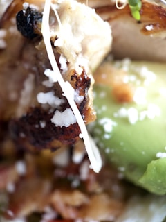 Close-up of various food ingredients including grilled meat with a charred surface, avocado, and shredded cheese.