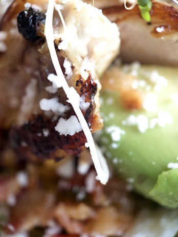 Close-up of various food ingredients including grilled meat with a charred surface, avocado, and shredded cheese.