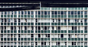 A repetitive grid pattern of windows on a building facade, featuring vertically and horizontally aligned green panels alternating with white framing. Most windows appear closed with some showing partially drawn curtains behind them.