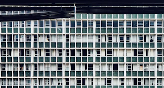 A repetitive grid pattern of windows on a building facade, featuring vertically and horizontally aligned green panels alternating with white framing. Most windows appear closed with some showing partially drawn curtains behind them.