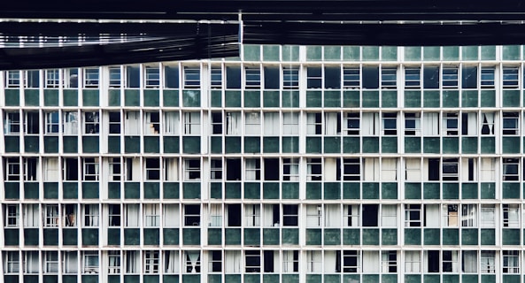 A repetitive grid pattern of windows on a building facade, featuring vertically and horizontally aligned green panels alternating with white framing. Most windows appear closed with some showing partially drawn curtains behind them.