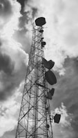 A tall metal communication tower reaches into a cloudy sky, with several large satellite dishes attached at various heights along its structure.