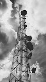 A tall metal communication tower reaches into a cloudy sky, with several large satellite dishes attached at various heights along its structure.