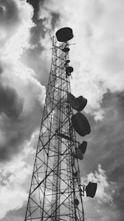 A tall metal communication tower reaches into a cloudy sky, with several large satellite dishes attached at various heights along its structure.
