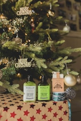 A beautifully decorated Christmas tree with gold and silver ornaments and 'Merry Christmas' signs. In front, there are skincare products labeled Noelle Australia, including body lotion and body wash, placed on a gift box wrapped in brown paper with red stars.