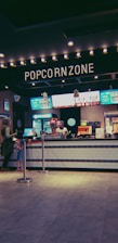 A concession stand at a movie theater with a prominent sign labeled 'Popcorn Zone.' The counter displays various snacks and a popcorn machine. Several digital menu boards feature colorful advertisements for food and beverages. Two people are standing in line to order.