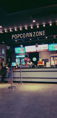 A concession stand at a movie theater with a prominent sign labeled 'Popcorn Zone.' The counter displays various snacks and a popcorn machine. Several digital menu boards feature colorful advertisements for food and beverages. Two people are standing in line to order.