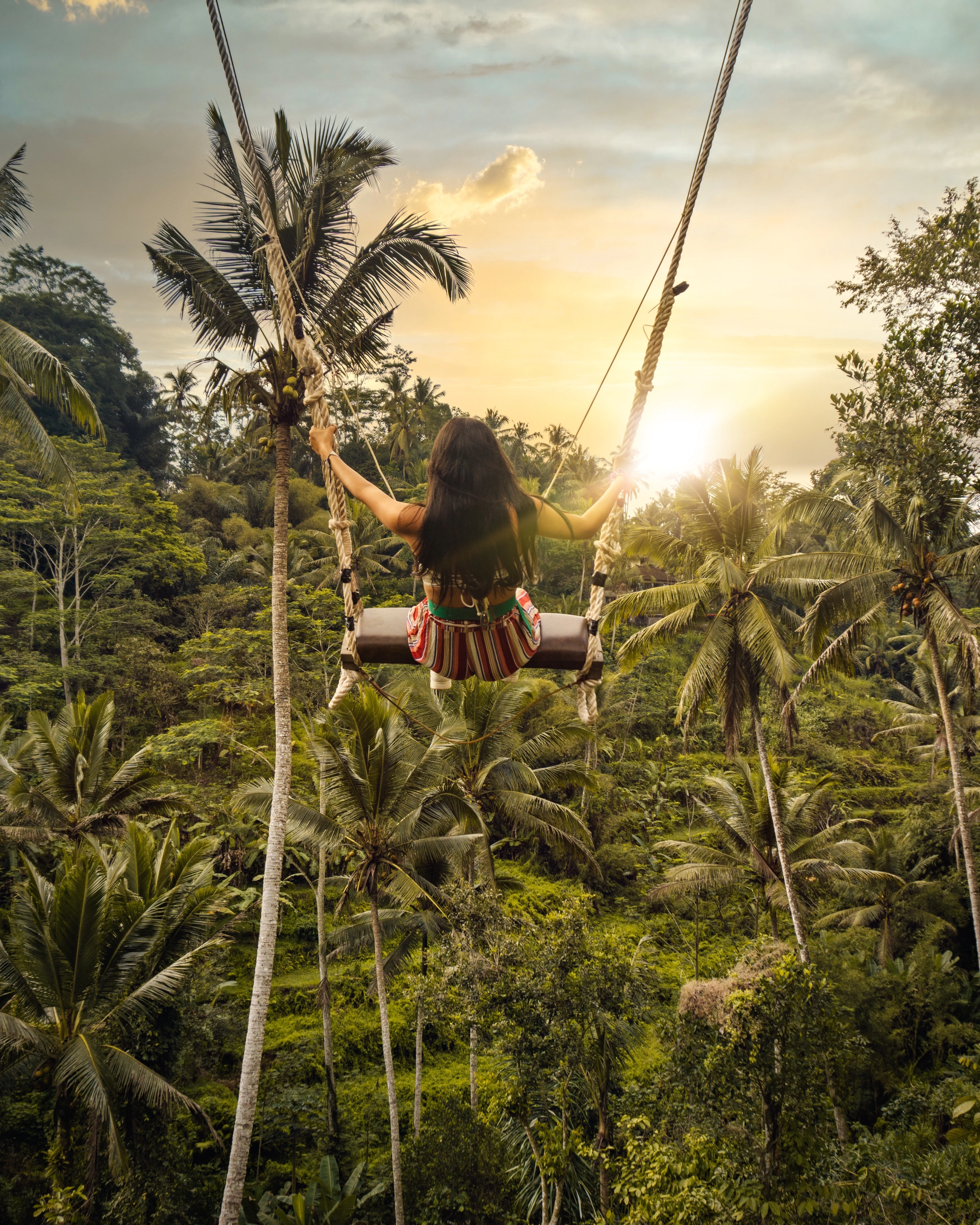 A woman on a jungle swing above the palm trees at sunset