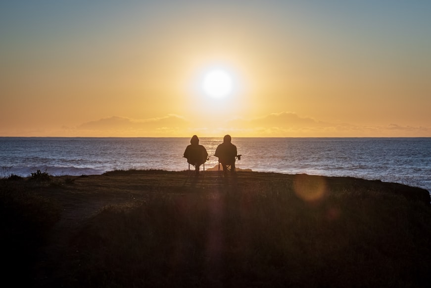 two silhouettes of people sitting on the edge of the water watching the sunset
