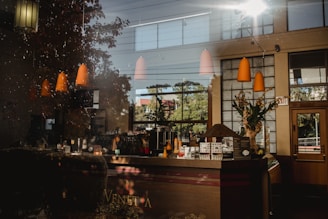 A cozy coffee shop interior with wooden counters and a variety of coffee-making equipment. Warm pendant lights hang from the ceiling, and large windows let in sunlight, casting shadows and reflections throughout the space. Decor includes potted plants and a display of menu items on the counter.