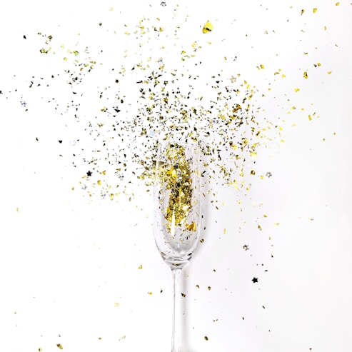Confetti explodes from a champagne glass, scattering gold and silver pieces against a white background.