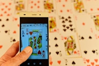 A hand is holding a smartphone displaying a close-up image of playing cards, specifically a King of Hearts and an Ace of Clubs. In the background, several playing cards are spread out, featuring various suits and numbers, slightly blurred for depth of field.