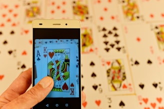 A hand is holding a smartphone displaying a close-up image of playing cards, specifically a King of Hearts and an Ace of Clubs. In the background, several playing cards are spread out, featuring various suits and numbers, slightly blurred for depth of field.