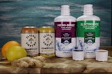 Two cans of Flying Embers hard kombucha in Ginger & Oak and Lemon Orchard flavors are placed next to two large bottles of supplements labeled Micro-Boost Humic Mineral Supplement and Electrolyze Fulvic Electrolyte Supplement. An orange, a lemon, and a piece of ginger rest on a wooden surface in front of a textured blue and green background.