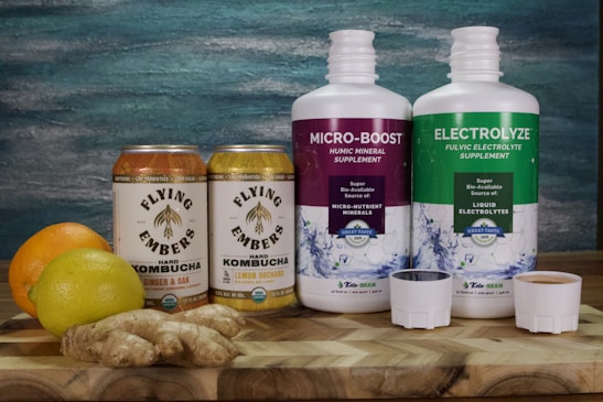 Two cans of Flying Embers hard kombucha in Ginger & Oak and Lemon Orchard flavors are placed next to two large bottles of supplements labeled Micro-Boost Humic Mineral Supplement and Electrolyze Fulvic Electrolyte Supplement. An orange, a lemon, and a piece of ginger rest on a wooden surface in front of a textured blue and green background.