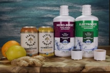 Two cans of Flying Embers hard kombucha in Ginger & Oak and Lemon Orchard flavors are placed next to two large bottles of supplements labeled Micro-Boost Humic Mineral Supplement and Electrolyze Fulvic Electrolyte Supplement. An orange, a lemon, and a piece of ginger rest on a wooden surface in front of a textured blue and green background.