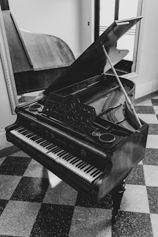 A grand piano with an open lid is positioned in a room with a checkered floor pattern. The intricate detailing on the music stand and the polished wood finish create a classic and elegant atmosphere. A mirror on the wall reflects the piano's shape, adding depth to the image.
