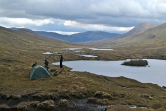 A remote and serene landscape with rolling hills and multiple small lakes. In the foreground, a group is camping with a green tent pitched near the water. The overcast sky adds a moody atmosphere while the distant mountains form a dramatic backdrop.