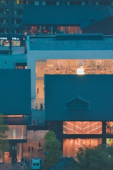 A modern building with a prominent glass facade showcasing a large illuminated logo. Inside, people are visible interacting in a bright, minimalist interior. Surrounding the central structure are traditional-style buildings with dark roofs, creating a contrast between modern and traditional architecture.