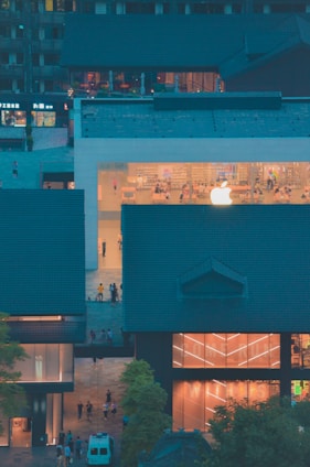A modern building with a prominent glass facade showcasing a large illuminated logo. Inside, people are visible interacting in a bright, minimalist interior. Surrounding the central structure are traditional-style buildings with dark roofs, creating a contrast between modern and traditional architecture.