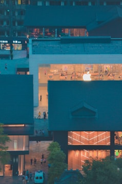 A modern building with a prominent glass facade showcasing a large illuminated logo. Inside, people are visible interacting in a bright, minimalist interior. Surrounding the central structure are traditional-style buildings with dark roofs, creating a contrast between modern and traditional architecture.