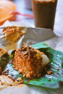 A serving of nasi lemak wrapped in banana leaves, garnished with crispy anchovies, sambal, and a piece of boiled egg. The food is placed on unwrapped brown paper, with a refreshing iced drink in the background.