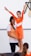A young girl in an orange gymnastics outfit stands with her arms raised, supported by an adult wearing a matching top. They are in an indoor setting with a white tiled wall in the background. Another child is partially visible in the foreground.