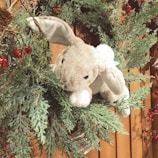 A plush bunny nestled among green pine branches adorned with small red berries and cones, against a wooden background. The bunny is soft and cuddly, with floppy ears and an inviting appearance.