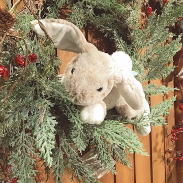 A plush bunny nestled among green pine branches adorned with small red berries and cones, against a wooden background. The bunny is soft and cuddly, with floppy ears and an inviting appearance.