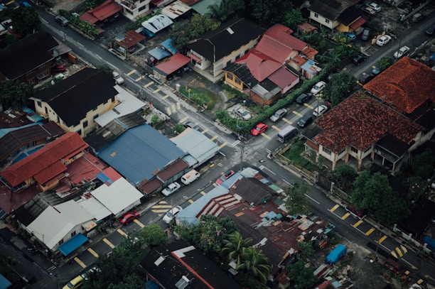 An aerial view of a residential neighborhood with a variety of houses featuring different colored roofs. Narrow streets intersect at right angles, lined with parked cars. Green trees and vegetation are visible throughout the area, creating a contrast with the urban infrastructure.