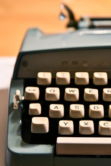 A close-up of an old-fashioned typewriter, focusing on the keys. The keys are arranged in a typical QWERTY layout and have a beige color. The typewriter itself is a teal or light blue color. The background is blurred, emphasizing the typewriter keys in the foreground.