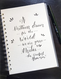 A spiral-bound dotted notebook lies open on a dark wooden surface. On the page, a motivational quote is written in an artistic, cursive style using black ink. A black pen rests next to the notebook.
