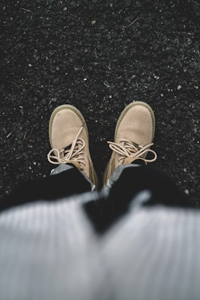 A pair of beige boots worn by a person standing on a rough, dark textured surface, possibly asphalt. The boots are laced up, and part of the person's clothing, including a sweater or jacket, is visible in the lower portion of the image.