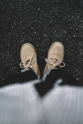 A pair of beige boots worn by a person standing on a rough, dark textured surface, possibly asphalt. The boots are laced up, and part of the person's clothing, including a sweater or jacket, is visible in the lower portion of the image.
