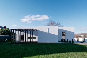 A modern, minimalist house with a white facade and large vertical wooden slats creating a covered area on one side. Manicured grass surrounds the building, and there is a small line of trees or shrubs planted near the entrance. The sky is clear with a few clouds, and a bare tree is visible in the background.