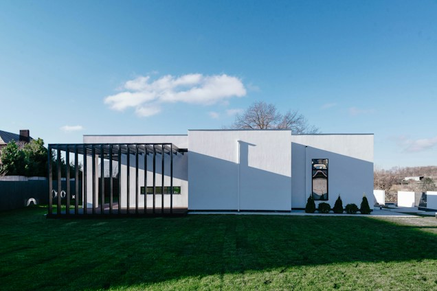 A modern, minimalist house with a white facade and large vertical wooden slats creating a covered area on one side. Manicured grass surrounds the building, and there is a small line of trees or shrubs planted near the entrance. The sky is clear with a few clouds, and a bare tree is visible in the background.