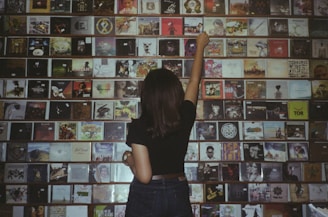 A person with long hair and a black shirt stands facing a wall filled with CDs. The CDs are displayed on shelves in rows, showcasing a variety of album covers, each with distinct artwork and colors. The person is reaching up with their left hand towards one of the CDs on the wall.