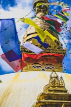 A large, golden stupa topped with colorful prayer flags. The stupa features a prominent pair of eyes, symbolizing wisdom and awareness, set against a sky with fluffy clouds. The surrounding flags add a sense of movement and vibrancy.