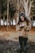 A young person with long curly hair holds a smartphone while standing outdoors in a forested area. They are wearing a brown hoodie and dark pants, with patterned rubber boots. The background features tall trees with soft sunlight filtering through.