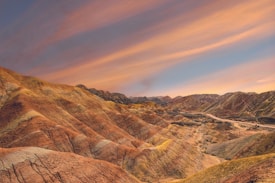 A striking landscape of rainbow-colored mountains featuring layers of red, orange, yellow, and green hues under a partially cloudy sky. The natural formations create a wave-like pattern, accentuated by the textured lines of the ridges.
